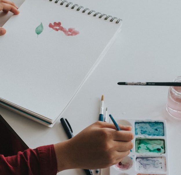Paint A hand holding a paintbrush above a palette with watercolors and a sketchbook nearby.